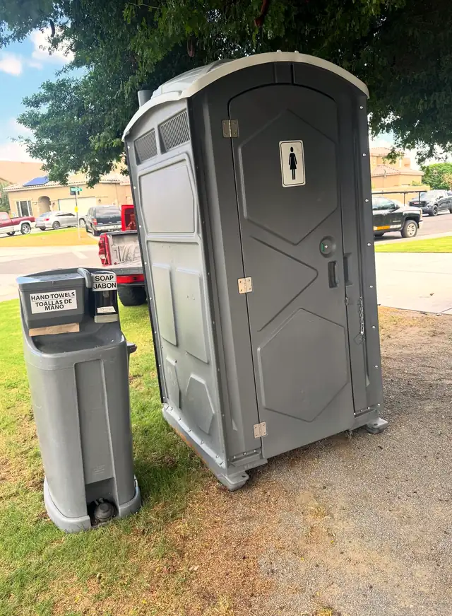 Gray portable toilet next to a freestanding handwashing station on a lawn